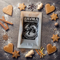 Beutel Hayma Neunerlei Pfefferkuchengewürz auf einem mit Mehl bestäubten Holztisch, ringsum Lebkuchen, Zimtstangen, Nelken, Ingwer, Muskatnuss und Orangenschale